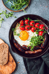 Breakfast time, fried egg with sausages and cherry tomatoes in a black iron pan, served microgreens.