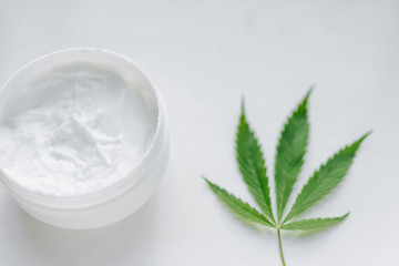Obraz na płótnie Canvas Open jar of hemp cream with cannabis leaf on white isolated background. Top view with moisturizing lotion from natural products containing CBD. Flat composition with marijuana and copy space