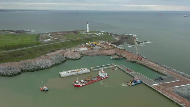 Aerial view of construction work on the new Aberdeen South Harbour at Nigg Bay on a cloudy day. Right to left circling shot around machinery with the lighthouse behind. Slow zoom in.