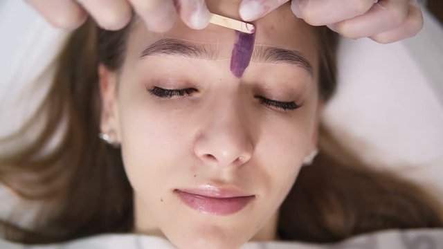 Young, caucasian girl is lying on couch during eyebrowes treatment at studio beauty, beautician depilating and shaping brows at beauty salon, removing the wax between the brows using a stick. Footage