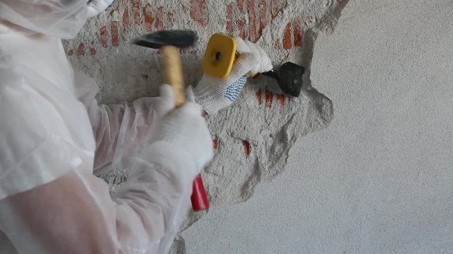 A man in equipment destroys a plastered wall with a chisel and hummer