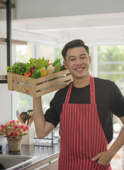 Young happy Asian man holding vegetable basket in kitchen room