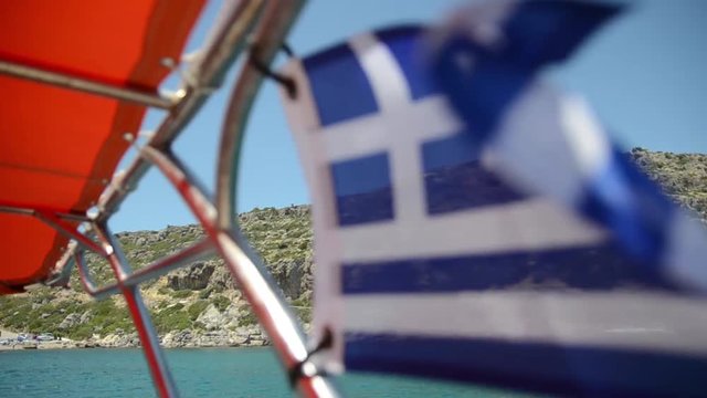 Flag of Greece flying in the wind on boat on sea, focused on island on background
