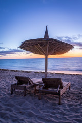 Anakao beach, Tulear, Madagascar.  Wooden reclining lounge chairs  with thatched roof umbrellas on a sandy beach with sunset over the ocean. 