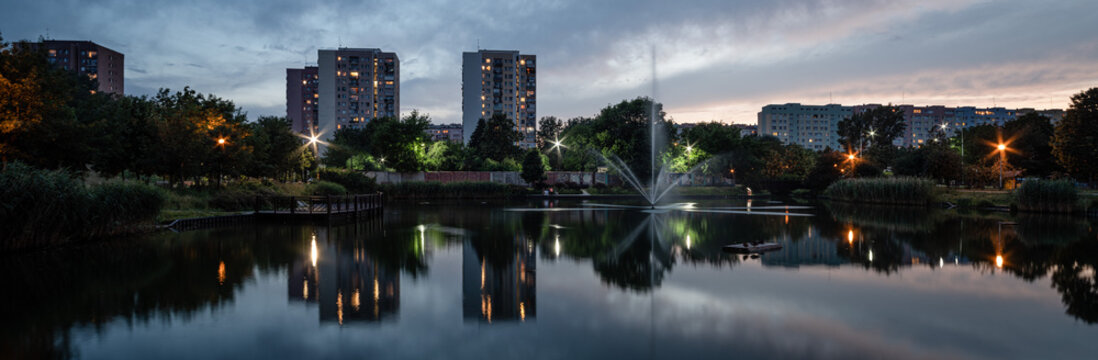 Urban evening scene. Apartment buildings stand among trees. A fountain on a city pond in the foreground.