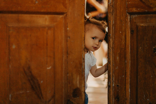 A Pretty Little Girl Hiding Behind A Large Wooden Door Looking In The Gap Between Them. Selective Focus, Noise Effect