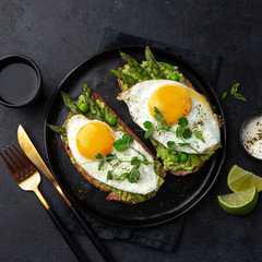toasts with avocado, asparagus and fried egg on black plate, top view, dark background,