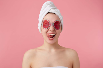 Close up of young funny lady with mask on eyes, after shower with a towel on her head, looks at the camera, shows tongue and stands over pink background.