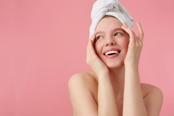 Studio photo of young happy woman after shower with a towel on her head, broadly smiles, looks away, touches face and smooth skin, stands over pink background.