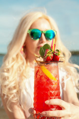 Beautiful blonde girl with a red beautiful cocktail in her hands by the sea / river