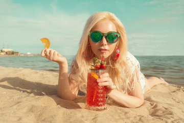 Beautiful blonde girl with a red beautiful cocktail in her hands by the sea / river