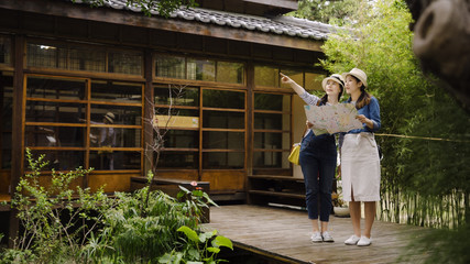 Attractive asian female traveler having conversation with best friend pointing finger discussing route. two girls searching location on paper map standing on wooden bridge out of japanese style house