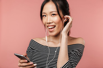 Photo of attractive asian woman dressed in basic wear holding smartphone and listening to music with earphones