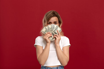 Portrait of a girl with curly blond hair dressed in a white t-shirt standing on a red background. Happy model hides behind dollars.