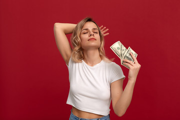 Portrait of a girl with curly blond hair dressed in a white t-shirt standing on a red background. Happy model .fans herself with a bundle of dollars with dreamy look.