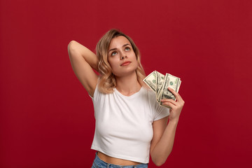 Portrait of a girl with curly blond hair dressed in a white t-shirt standing on a red background. Happy model .fans herself with a bundle of dollars with dreamy look.