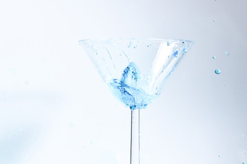 Cocktail with blue liquid in glass. Glass with blue water pouring with liquid with splashes and drops. Martini glass filling with alcohol with splashes on white background. Refreshing drink concept.