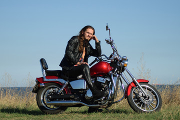 Plakat Handsome long-haired biker in the leather on red motorcycle against field and seashore. Brutal man in leather