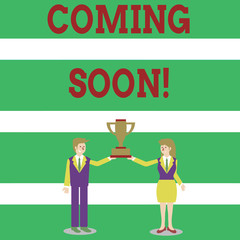 Writing note showing Coming Soon. Business concept for event or action that will happen after really short time Man and Woman Business Suit Holding Championship Trophy Cup