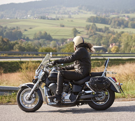 Fototapeta na wymiar Side view of bearded motorcyclist in helmet and black leather clothing riding bike along sharp turn of empty road on bright summer day, on misty background of rural landscape and distant green hills.