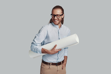 Young architect. Handsome young man carrying blueprint and smiling while standing against grey background