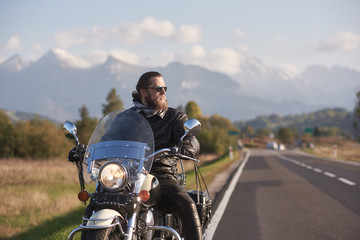 Fototapeta na wymiar Portrait of handsome bearded biker in black leather jacket and sunglasses holding motorbike handles on country roadside on blurred background of green landscape, distant white mountain peaks.