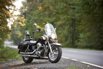 Black shiny powerful cruiser motorcycle parked on roadside on background of green trees on sunny...