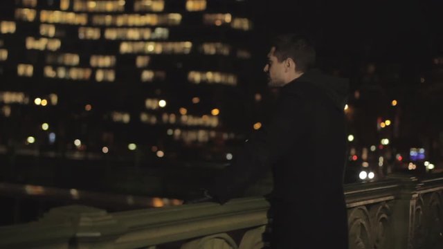 Depressed handsome man with beard and brown hair leaning on a railing of the bridge, thinking sadly to his ex girlfriend on blurred city colorful lights background. Medium Shot.