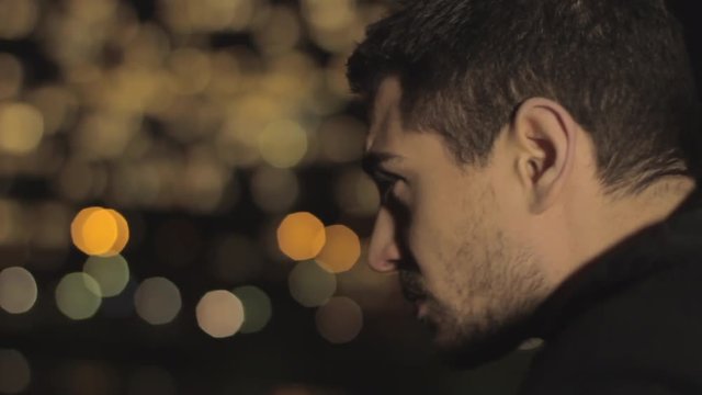 Sad handsome man with beard and brown hair leaning on a railing of the bridge, thinking sadly to his ex girlfriend on blurred city colorful lights background.