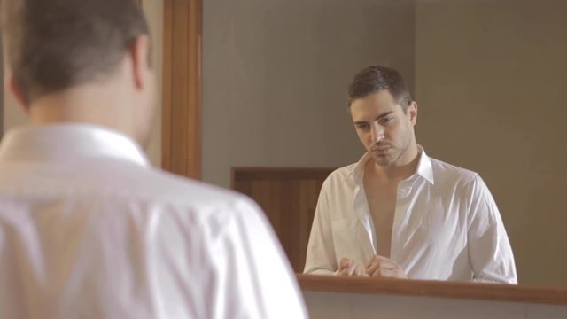 Handsome young man with a beard dressing with a white shirt and looking at himself in the mirror, thoughtful, sad and alone. Anxiety, breaking, argument, loneliness, sadness, depression, memories.