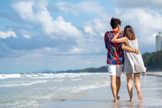 Back Rear View Of Young Adult Asian Couple Men And Women Enjoy Romantic Holiday Vacation Travel Holding Hands And Walking Together On The Island Beach And Sea In Summer With Blue Sky And White Clouds