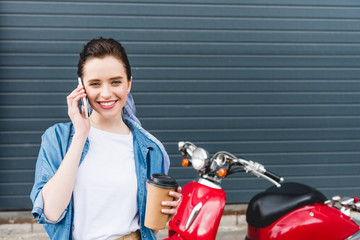 front view of beautiful girl standing near red scooter, holding paper cup with coffee and talking on smartphone