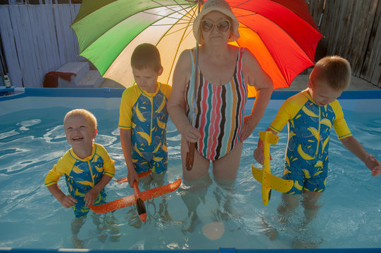 Elderly woman in striped swimsuit, sunglasses and elegant hat with rainbow color umbrella standing in pool. Children in swimwear swim next to their grandmother and play with bright plastic airplanes