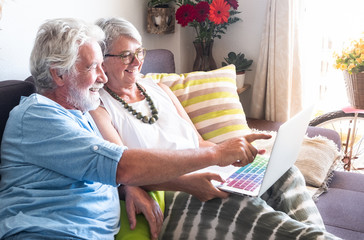 Couple of senior people laugh looking something funny in the laptop. Technology for old man and woman sitting on sofa with striped pillow