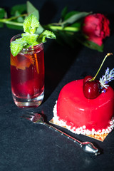 The unlearned cake covered with red icing lies on a dark background, next to a currant compote in a transparent glass, and there is a rose in the background. 
