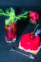 The unlearned cake covered with red icing lies on a dark background, next to a currant compote in a transparent glass, and there is a rose in the background. 