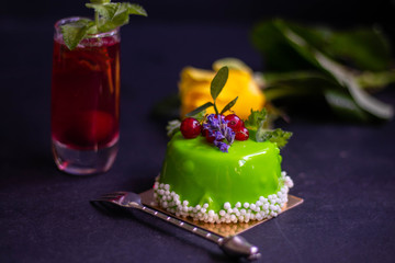 Unquoted cake in green glaze with currant berries stands on a dark background. Next to a glass of compote and cubes of cold, wet ice. 