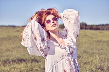 Fashion portrait. Beautiful woman in stylish dress walking at sunny grass field relax. Young romantic happy redhead Model girl enjoy summer nature in sunlight, fashionable outfit. Outdoor authentic