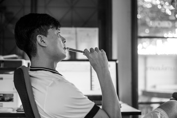 Closeup monochrome portrait Asian thoughtful man holds a pencil and thinking hard on his work with blurred bright screen of desktop computer in background