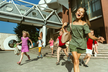 Happy kids playing at city's street in sunny summer's day in front of modern building. Group of happy childrens or teenagers having fun together. Concept of friendship, childhood, summer, holidays.