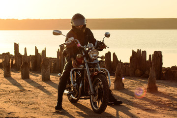 biker in the leather and helm on the red motorcycle against field and seashore. Brutal man in leather at sunset
