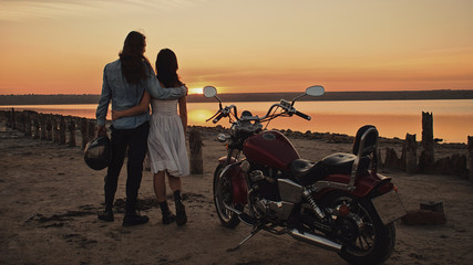 Romantic biker couple with red motorcycle. Handsome bearded long-haired man and attractive woman outdoors against motorcycle and sunset, sunrise