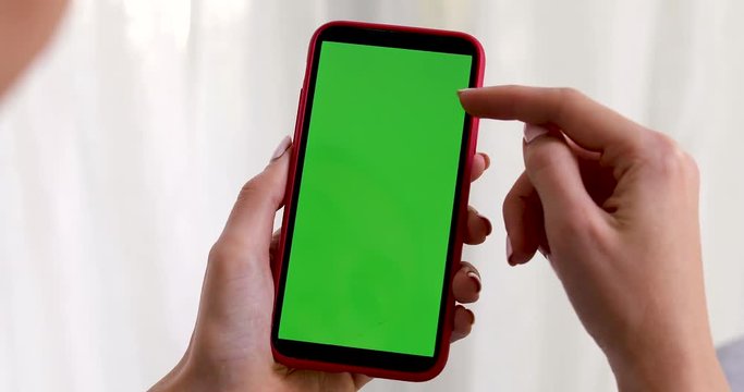 Female hand with a smartphone with a green screen on a white background curtains. Woman points her finger and touches the screen with her finger
