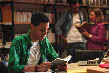Young male student study in the library reading, while his friend searching the book on the bookshelf behind him.