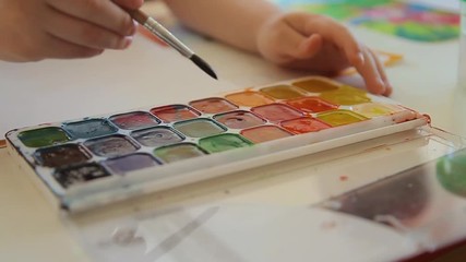 A child with a brush to paint selects the color of the paint. Set of watercolor paints of different colors. The boy takes the orange paint with a brush. Artist's palette, close-up. Drawing tools.