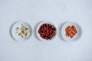 Halves of berries dark red cherries, separated bones and branches of cherries in different white bowls on white background