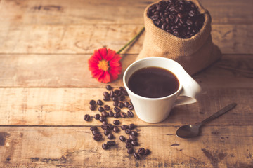 Black coffee In the cup white have roasted coffee beans are In a cloth bag sack and on table with, still have red flowers and stainless steel spoon all placed on a wooden table.