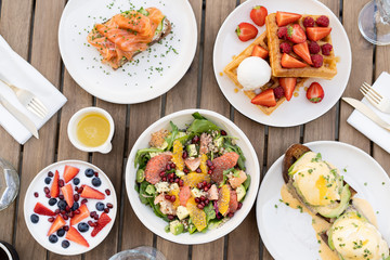 Stunning breakfast or lunch in open air in a cozy summer cafe. Poached eggs with Dutch sauce and avacado, coffee, bottle of water with mint, granola with fruit, waffles with ice cream, salad and chia