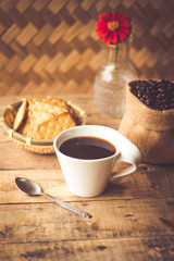 Black coffee In the cup white have roasted coffee beans are In a cloth bag sack butter cracker In the basket weave bamboo, still have red flowers and stainless steel spoon all placed on a wooden table