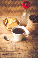 Black coffee In the cup white have roasted coffee beans are In a cloth bag sack butter cracker In the basket weave bamboo, still have red flowers and stainless steel spoon all placed on a wooden table